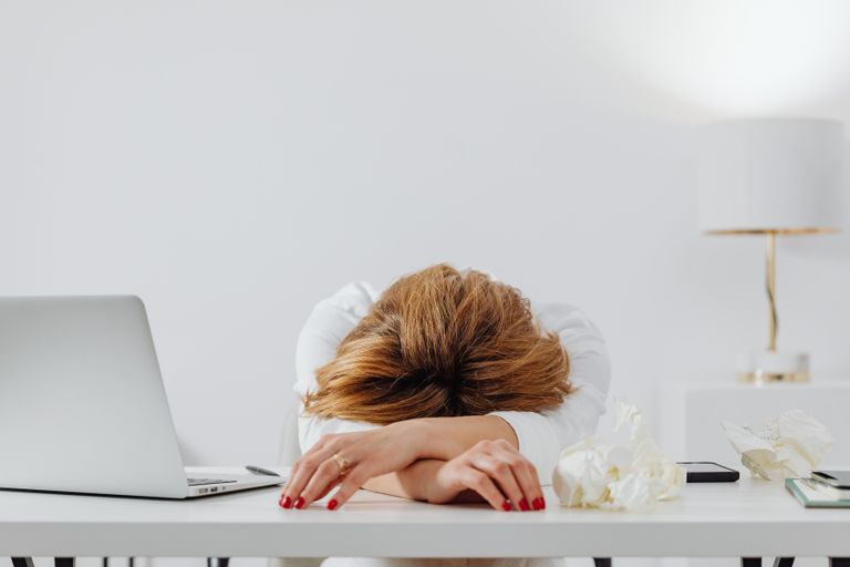 Tired woman resting head on arms at desk with laptop, showing fatigue in modern office.